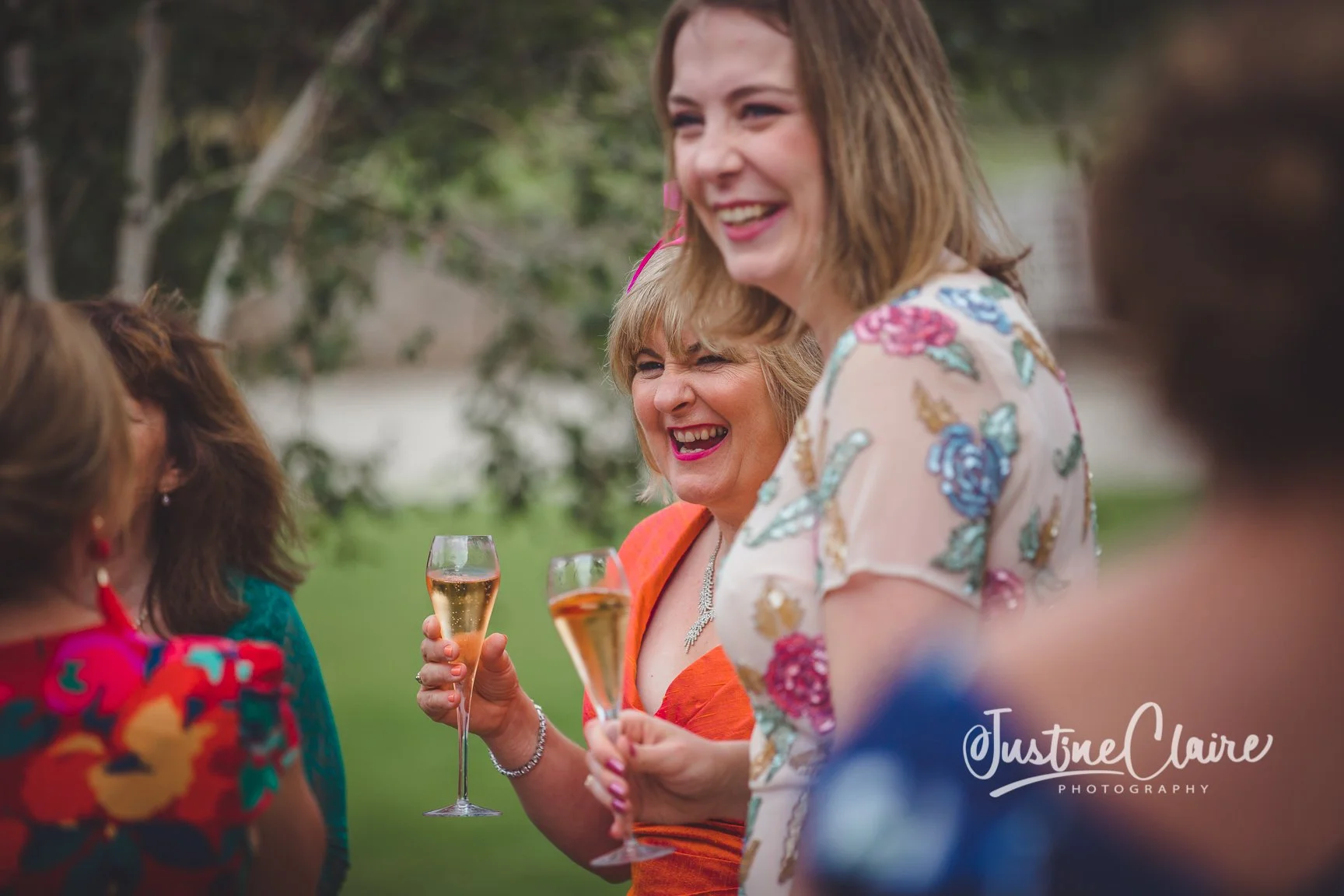 Women at a celebration outdoors holding glasses of champagne, smiling and laughing.