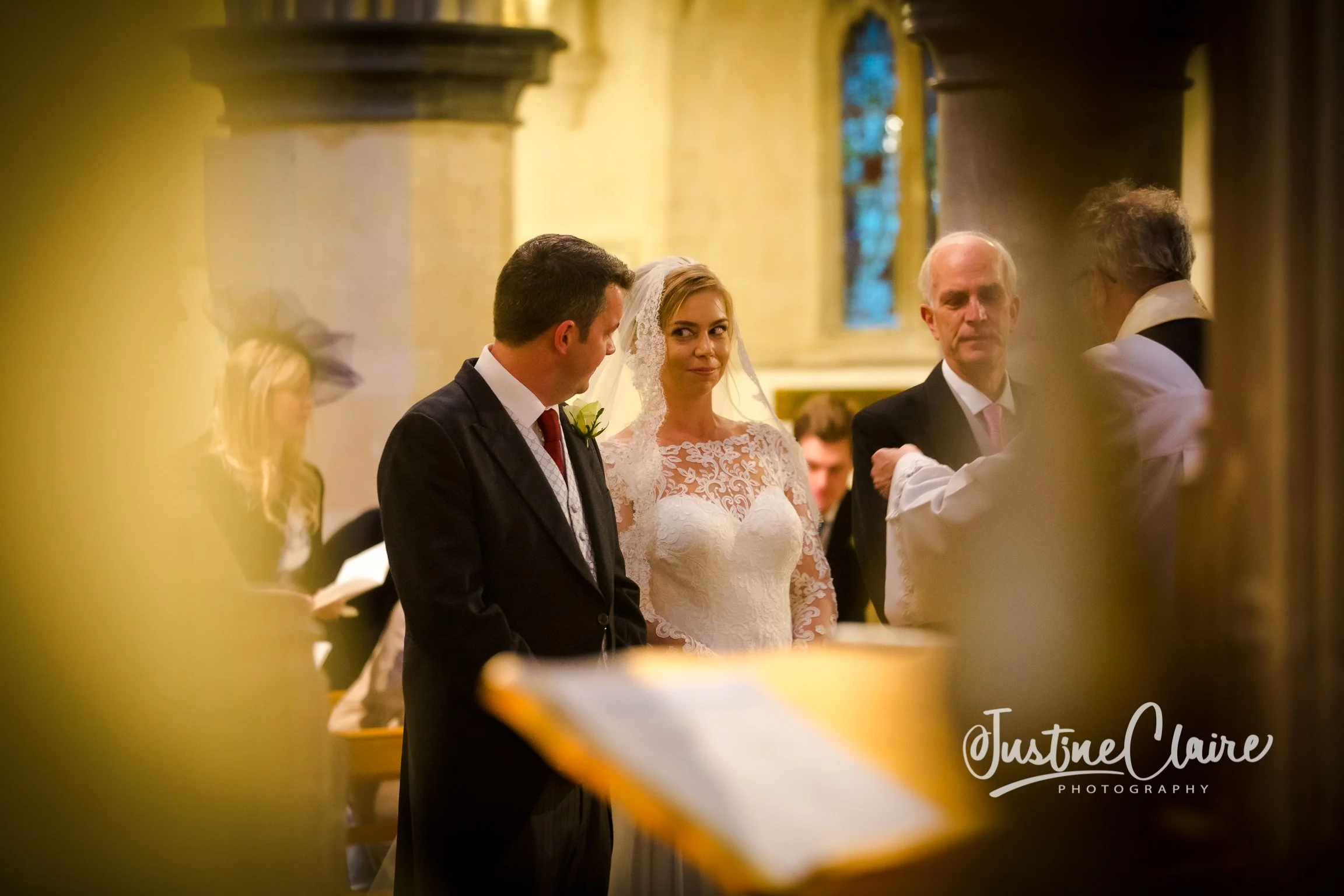 A wedding ceremony inside a church with a bride and groom standing before a priest. The bride wears a white lace wedding gown and veil, and the groom is in a dark suit with a white shirt and tie. Other guests are seen in the background.