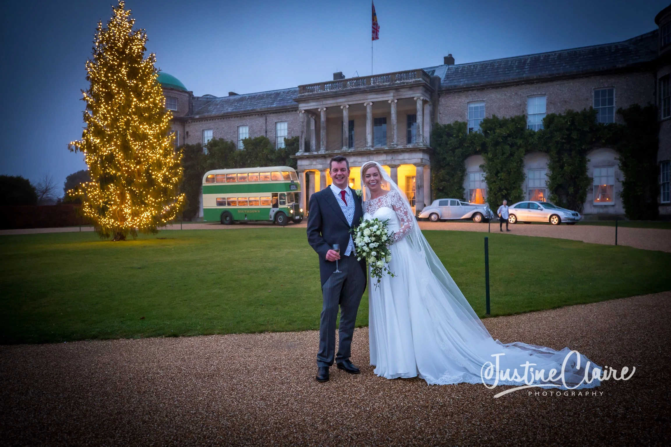 A newlywed couple standing outside a grand building with illuminated windows and a large Christmas tree decorated with yellow lights. The groom is in a suit holding a champagne glass, and the bride is in a white wedding dress holding a bouquet, smili