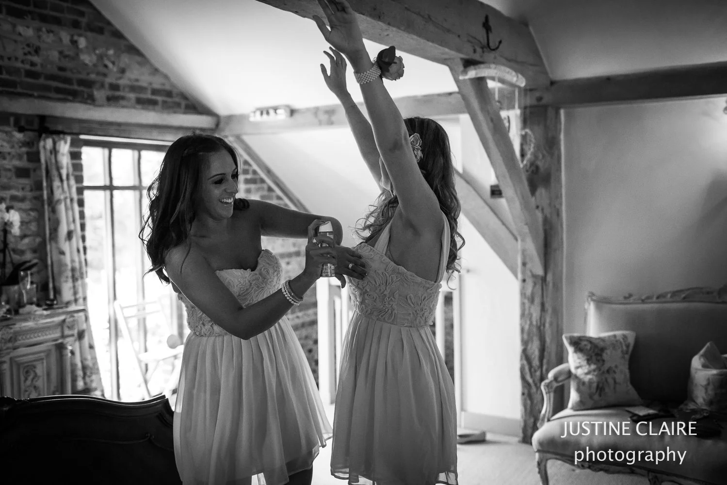 Two women in dresses smiling in a rustic room, one helping the other with a spray bottle. The woman on the right is raising her arms, and a camera is visible. The room has a sloped ceiling, brick wall, window, and decorative furniture.