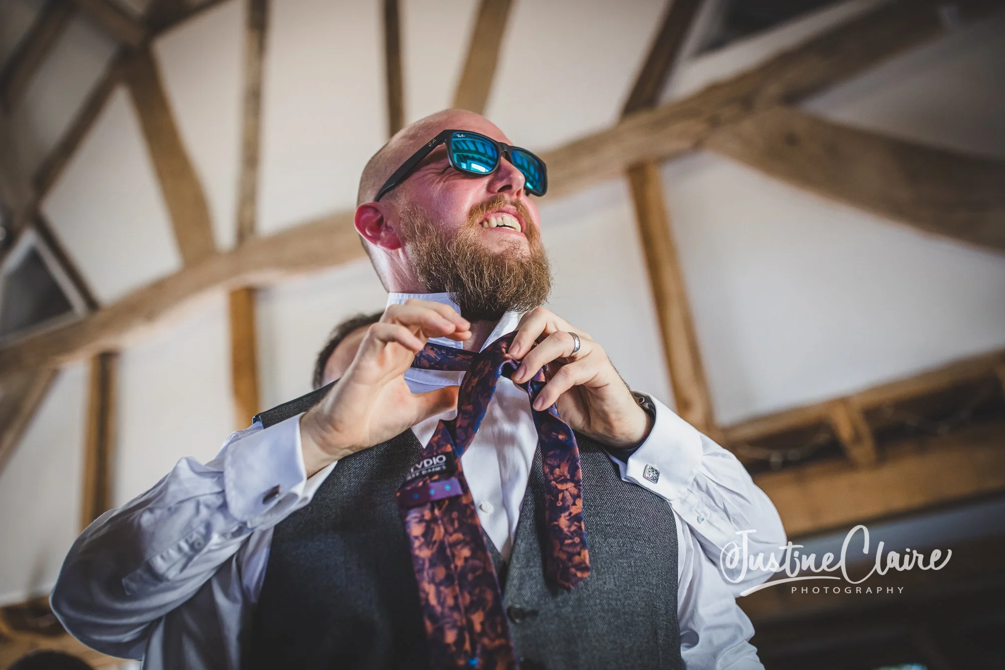 A man with a beard and sunglasses is adjusting a tie, wearing a white shirt and a dark vest, inside a wooden structure.