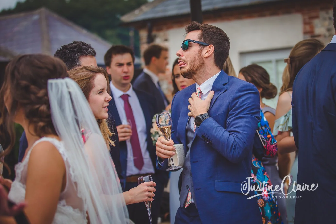 Group of wedding guests socializing outdoors, with a man wearing sunglasses and a blue suit, and a woman with a veil and a white dress, holding glasses of champagne.