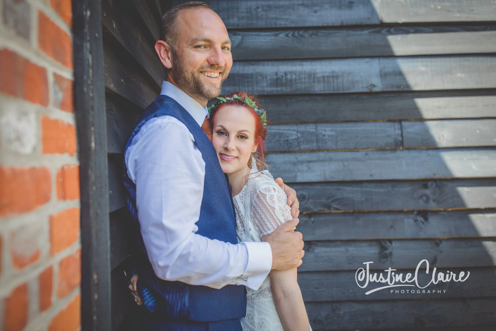A happy couple dressed in wedding attire, standing close together outdoors against a dark wooden wall, with the groom smiling at the camera and the bride resting her head on his shoulder.