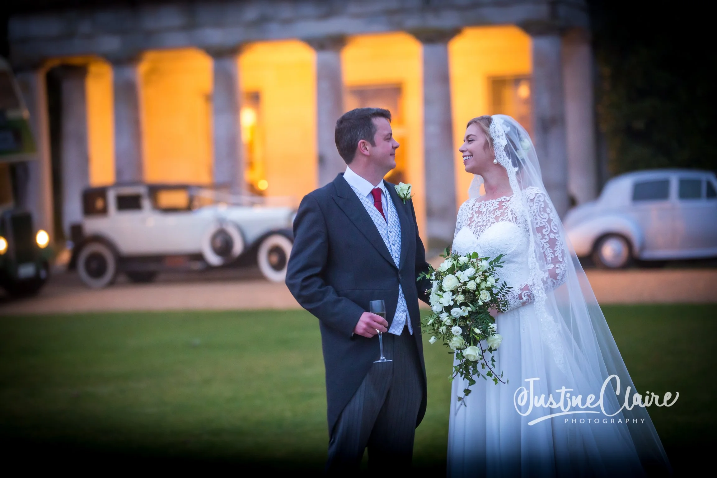 A bride and groom standing outdoors during their wedding, smiling at each other, with vintage cars and a warmly lit building in the background.