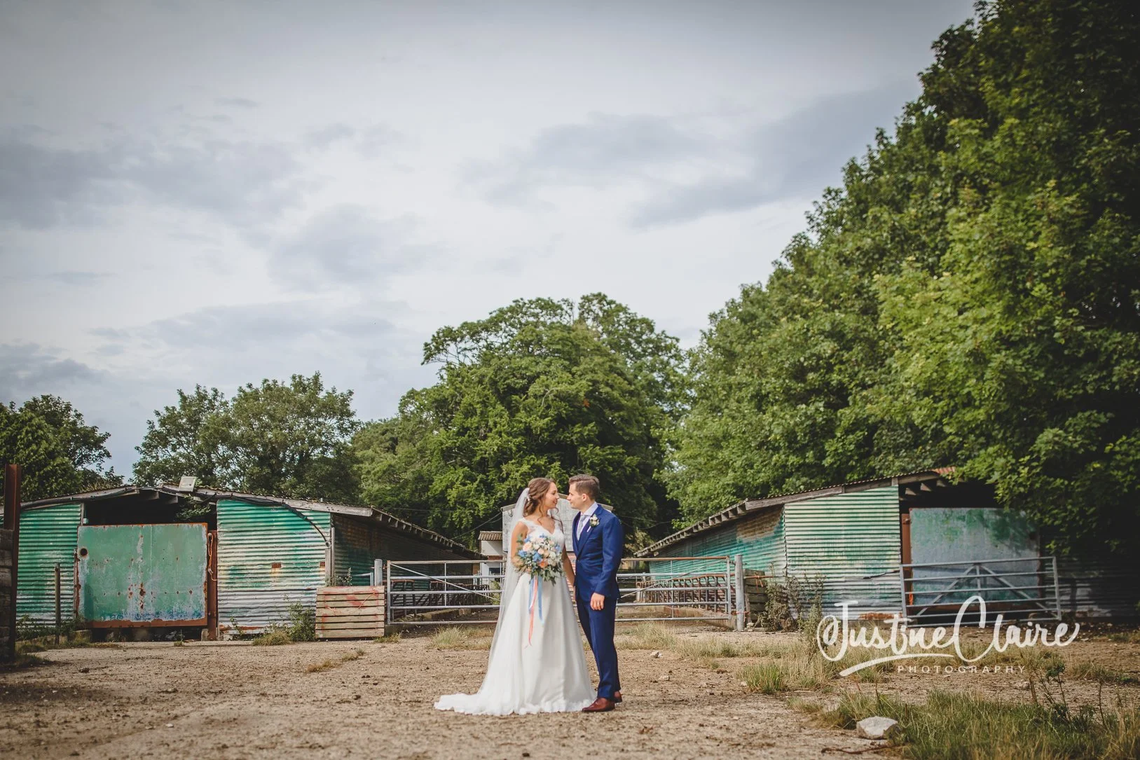 Bride and groom standing close together outdoors on a dirt path, with green trees and metal sheds in the background, exchanging looks on their wedding day.