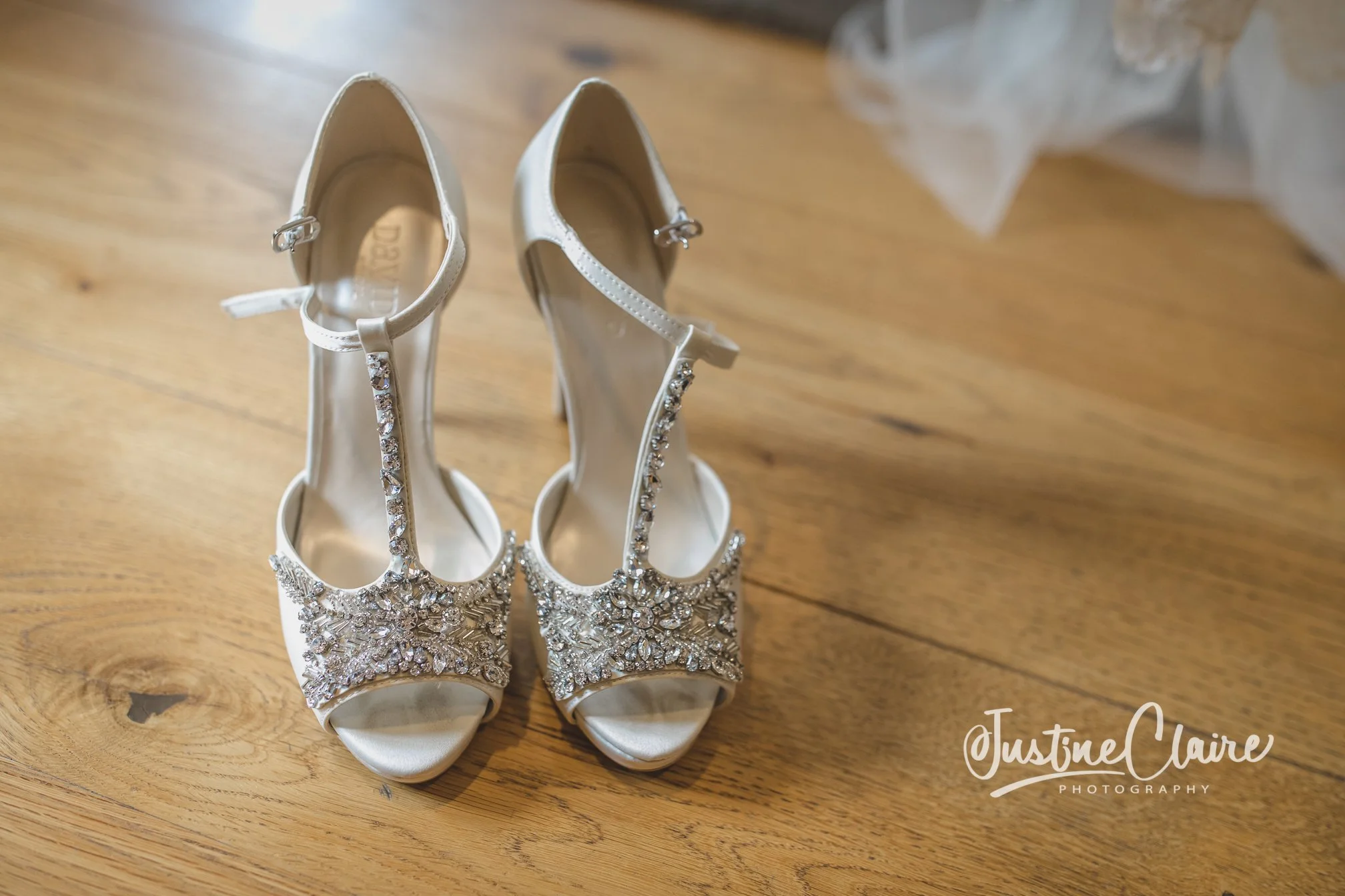 A pair of white wedding high-heeled shoes with jeweled embellishments on a wooden floor. farbridge wedding photography wedding shoes for farbridge