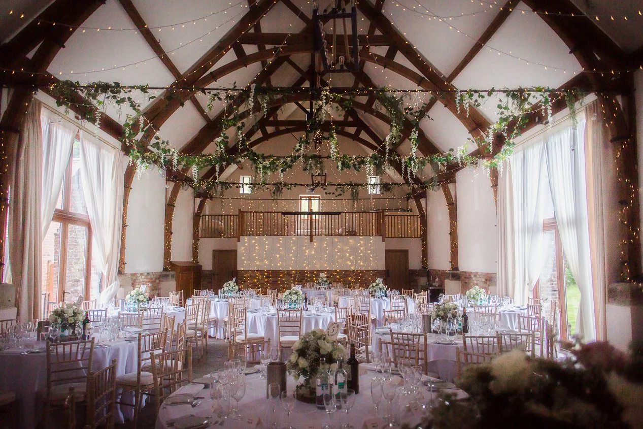 Decorated banquet hall with white tablecloths, floral centerpieces, and string lights hanging from wooden beams.