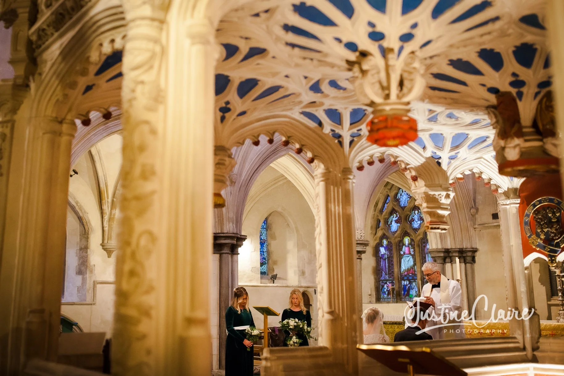 Inside a church with ornate architecture, two women in dark dresses holding bouquets, a man in white shirt and black bow tie reading from a paper, a bride with veil sitting, and stained glass windows in the background.