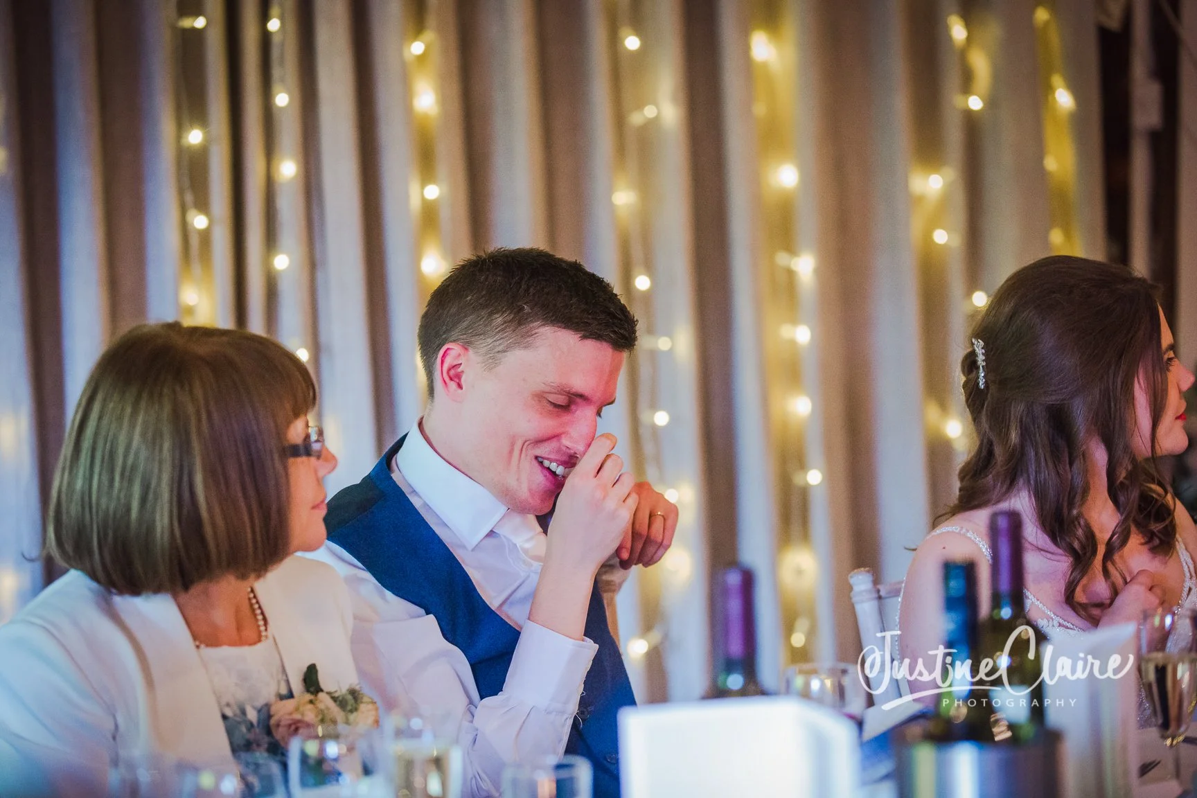 A man with short dark hair, wearing a white shirt and blue vest, is sitting at a decorated table with women, one with glasses and brown hair, and the other with long brown hair and a hair clip, in front of a backdrop of vertical lights. He appears to