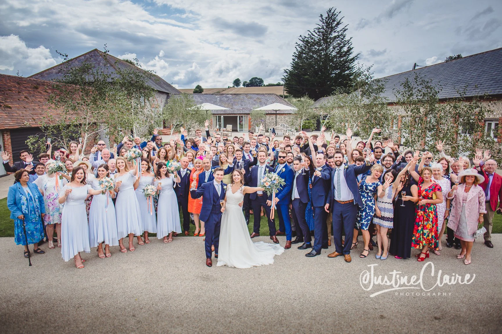 Group wedding photo outdoors featuring the bride and groom surrounded by wedding guests on a cloudy day, with houses and trees in the background.