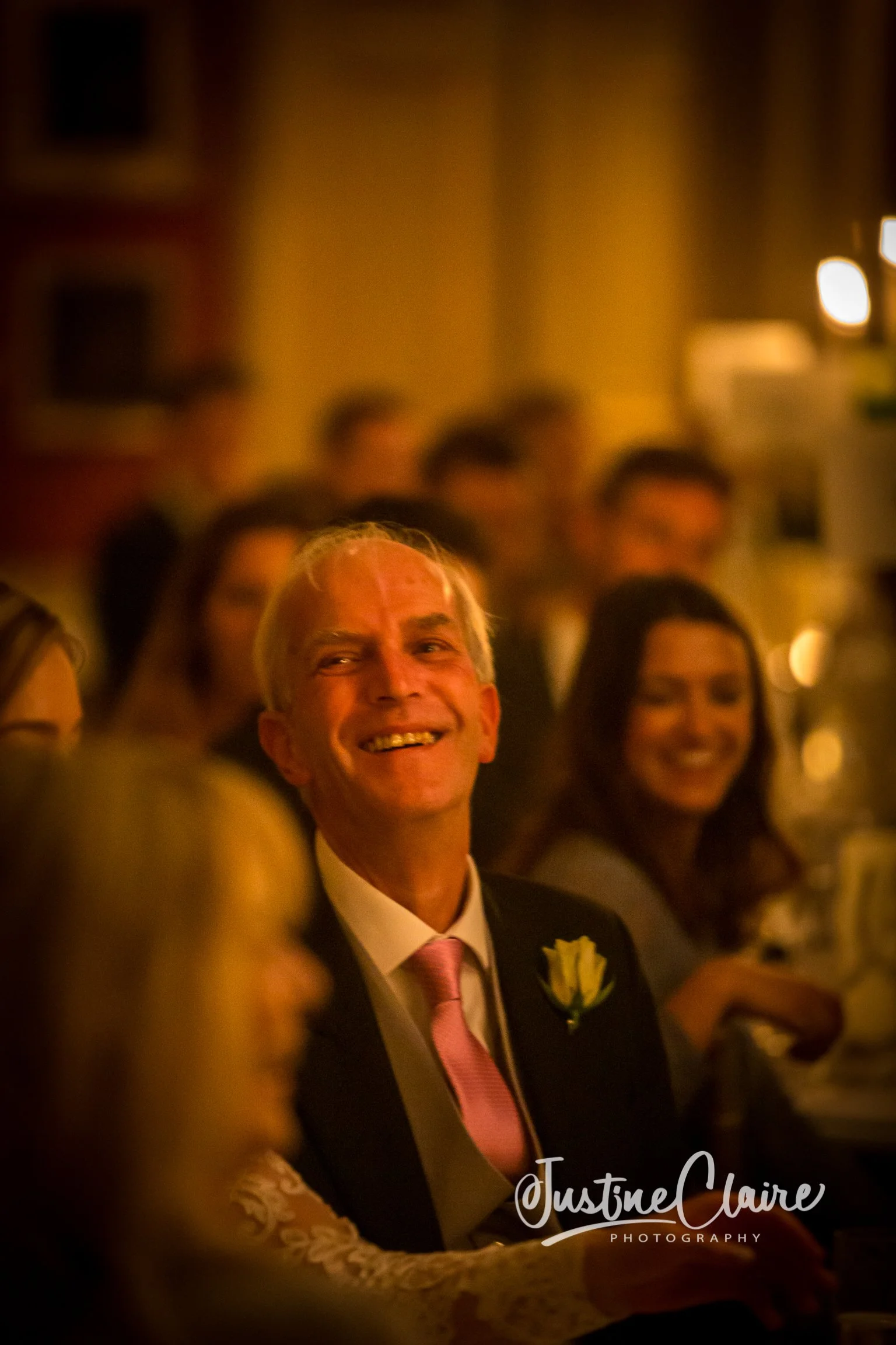 A man in a suit with a pink tie and a white flower boutonniere smiling at a formal event, with a woman seated behind him also smiling.