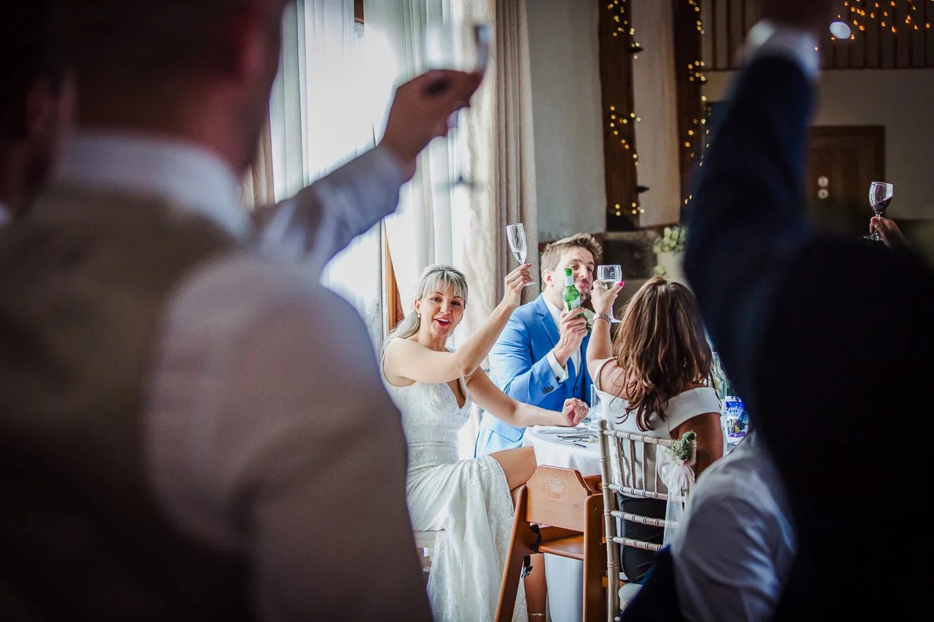 Woman in white dress and man in blue suit celebrating with champagne at a wedding reception, surrounded by guests raising glasses.