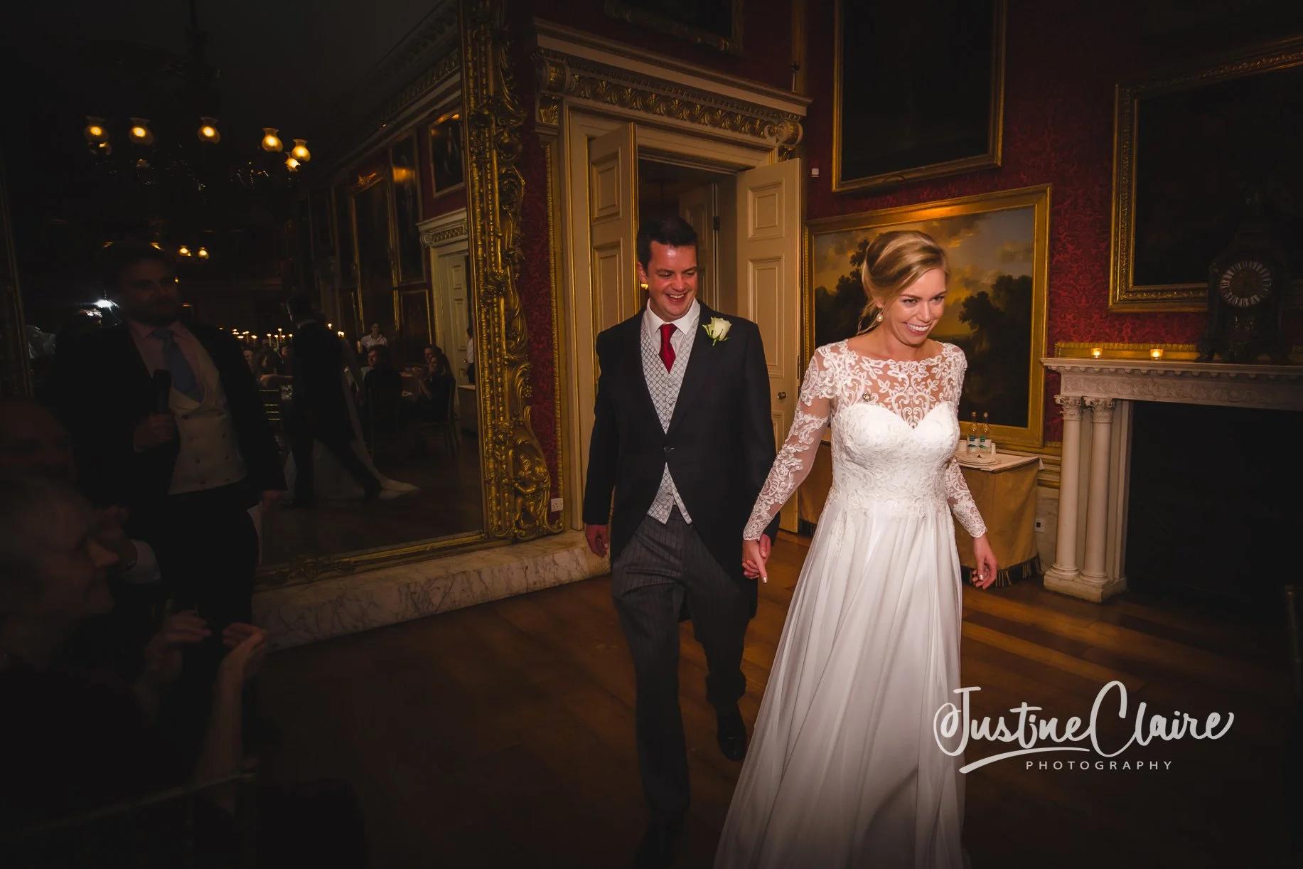 The bride and groom hold hands and walk into a wedding reception in an elegant, ornately decorated room with gold accents, paintings, and a fireplace, smiling happily.