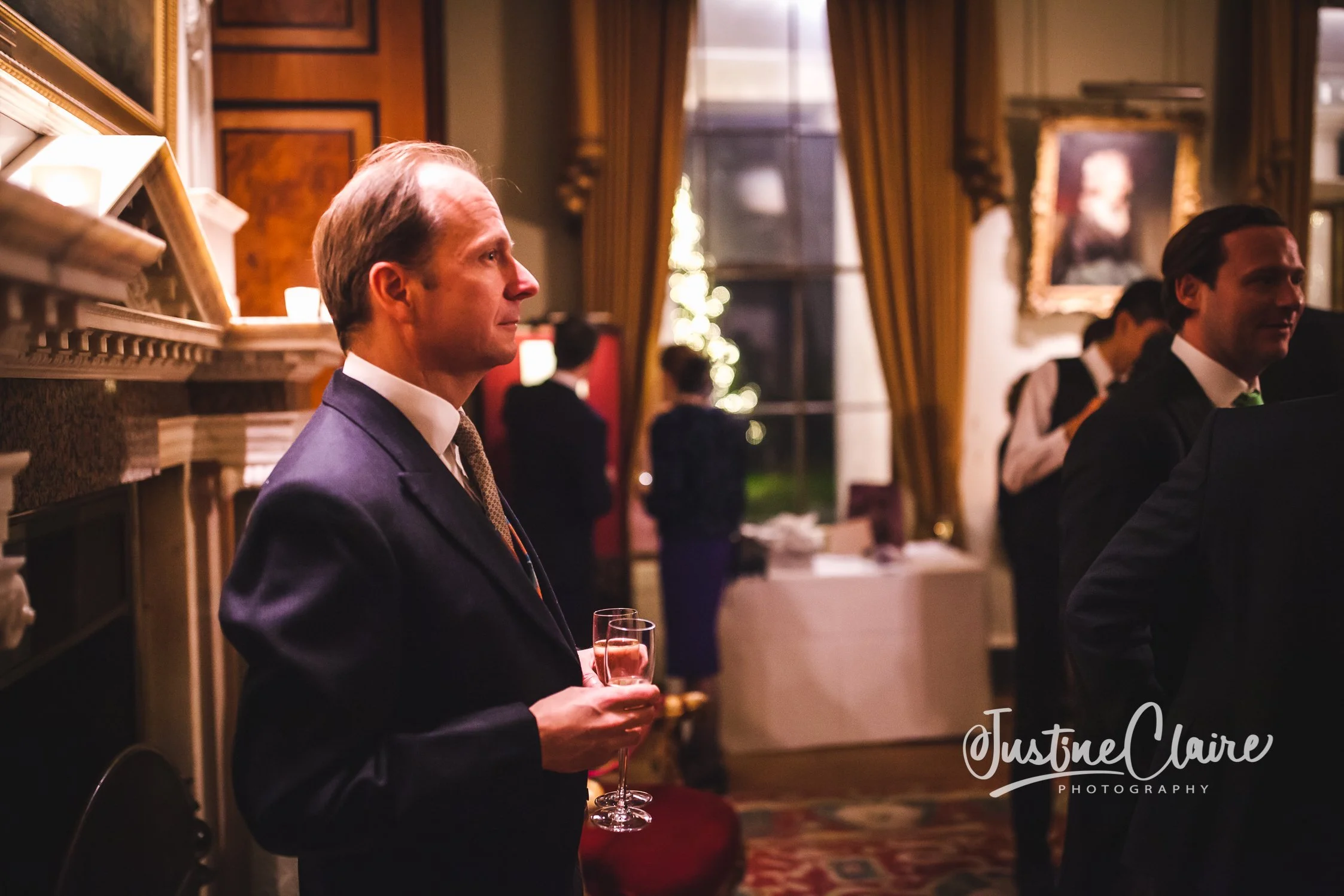 A man in a suit holding a glass at an indoor event with warm lighting, decorated with curtains, framed portraits, and festive lighting, during the evening.
