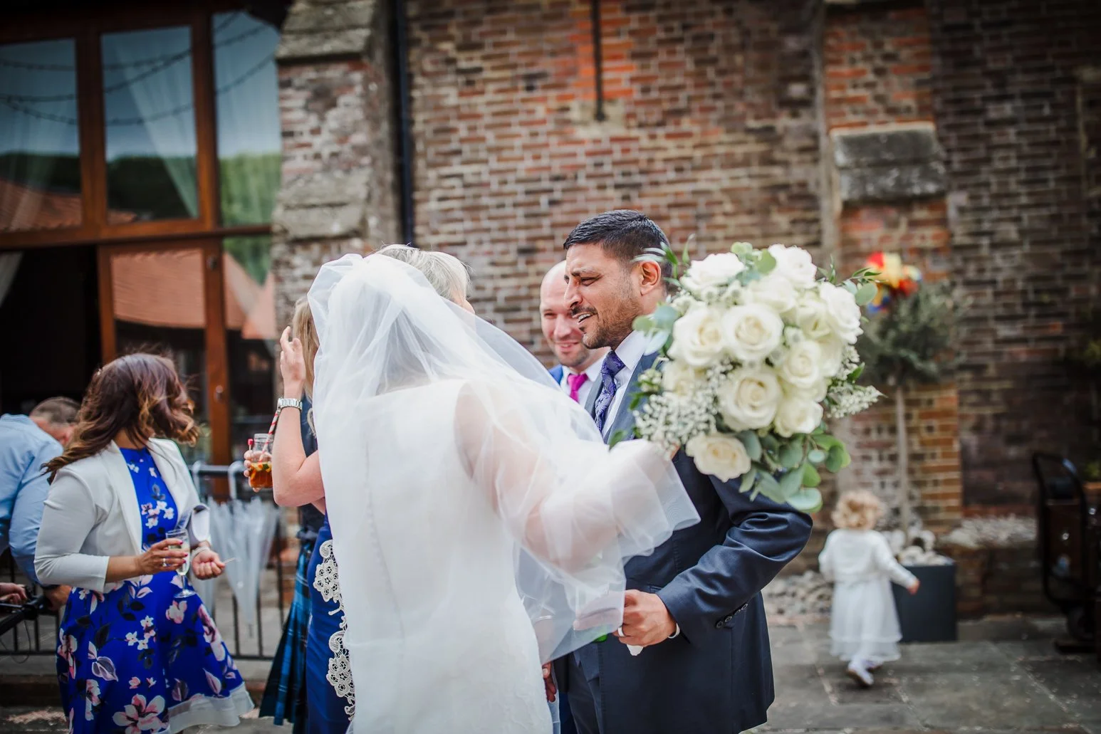 A bride and groom share an emotional moment during their wedding ceremony outdoors, with the bride holding a large bouquet of white roses, and guests in the background.