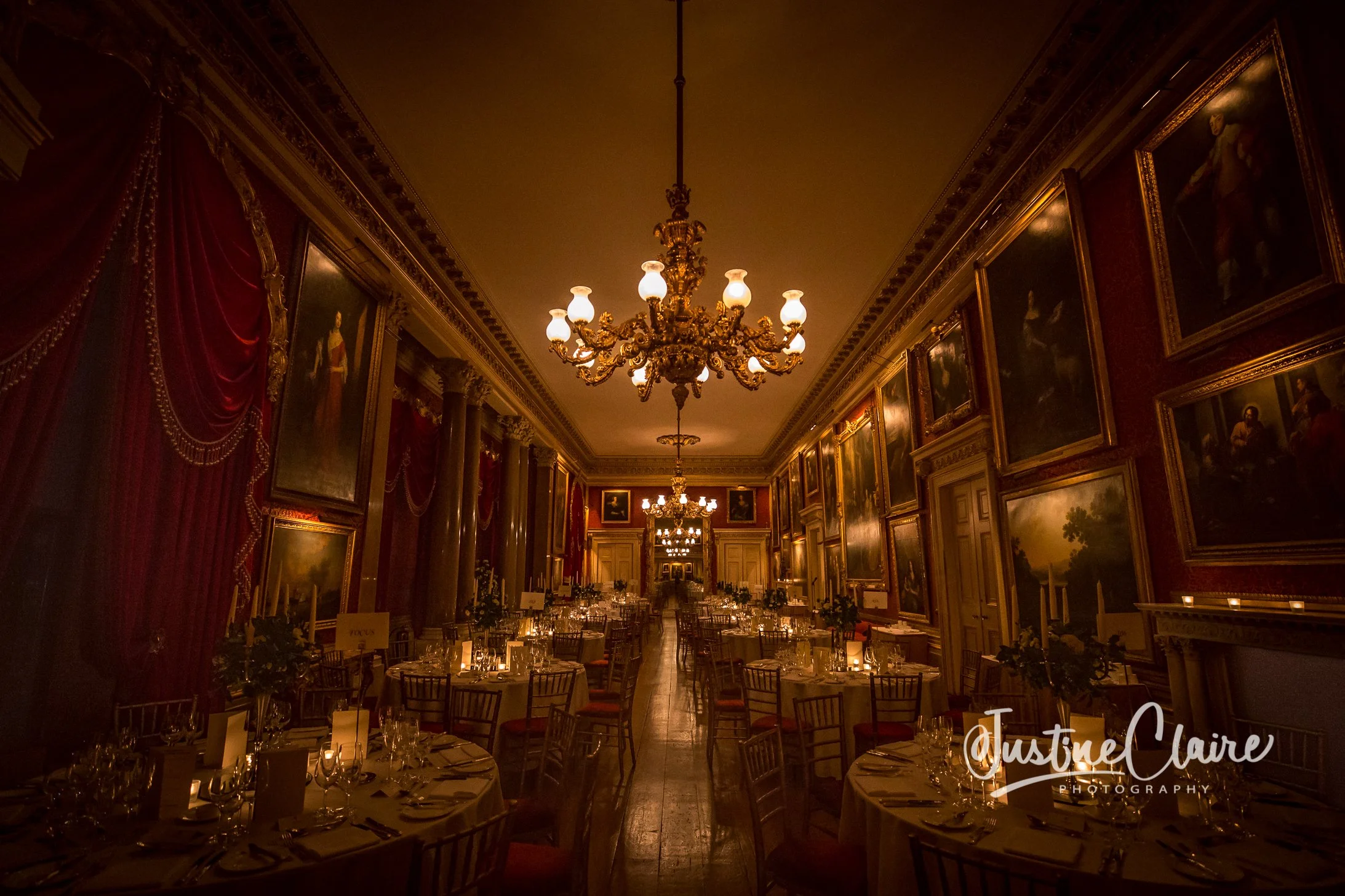 Elegant dining room with round tables, candlelit settings, ornate chandeliers, classical paintings, and red velvet curtains.