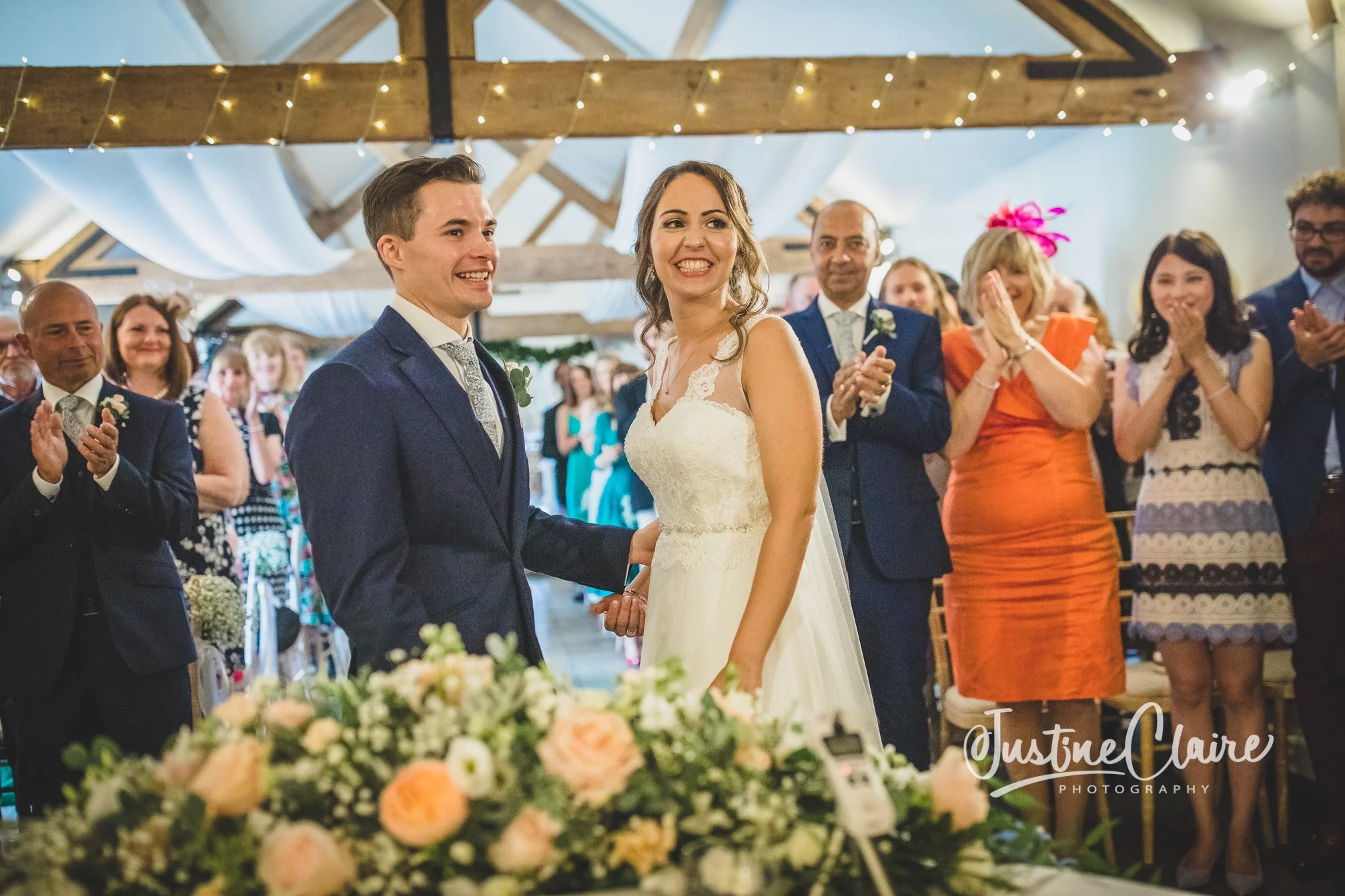 Bride and groom holding hands and smiling at their wedding ceremony, with guests clapping and celebrating in a decorated indoor venue. farbridge wedding photography