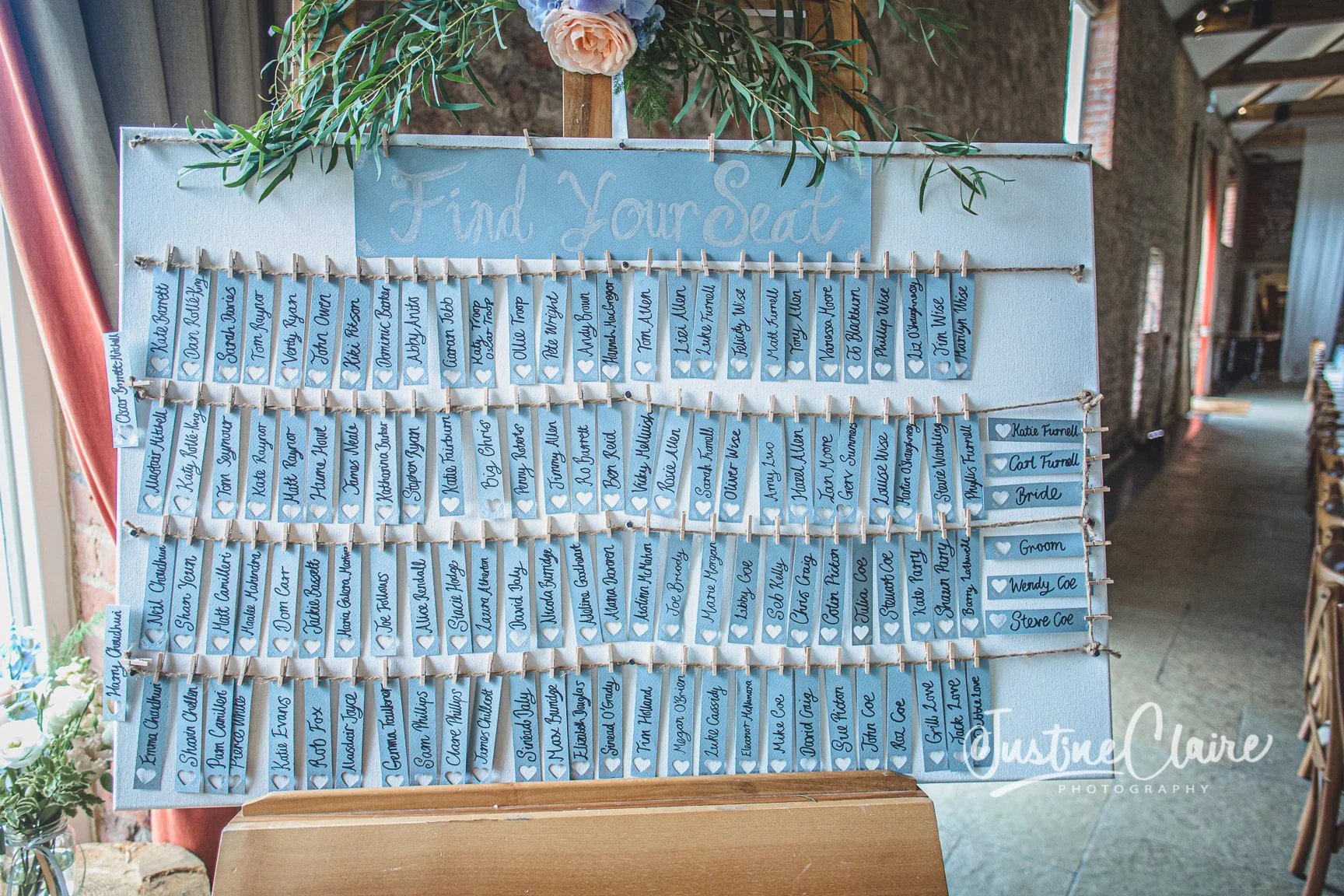 Wedding guest seating chart with blue tags displaying guest names, organized on ropes. The top has a sign that reads 'Find Your Seat' and is decorated with flowers and greenery.