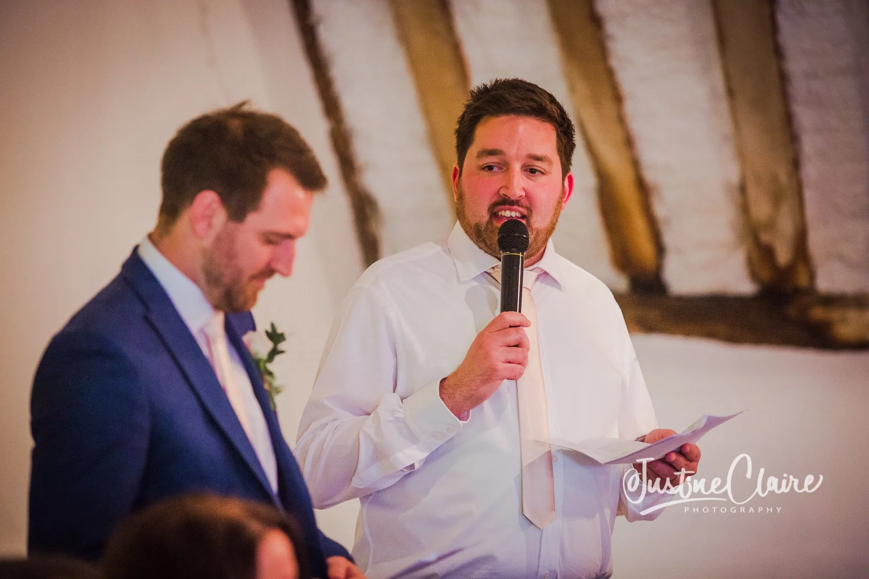 A man in a white shirt and tie is giving a speech at a wedding, holding a microphone in his right hand and reading from notes in his left hand, while another man in a blue suit stands nearby, both in a rustic venue with wooden beams.