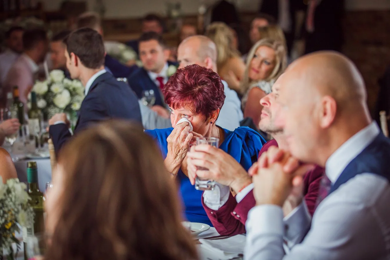 A woman with red hair in a blue dress is wiping tears from her eyes while holding a glass at a formal dinner gathering.
