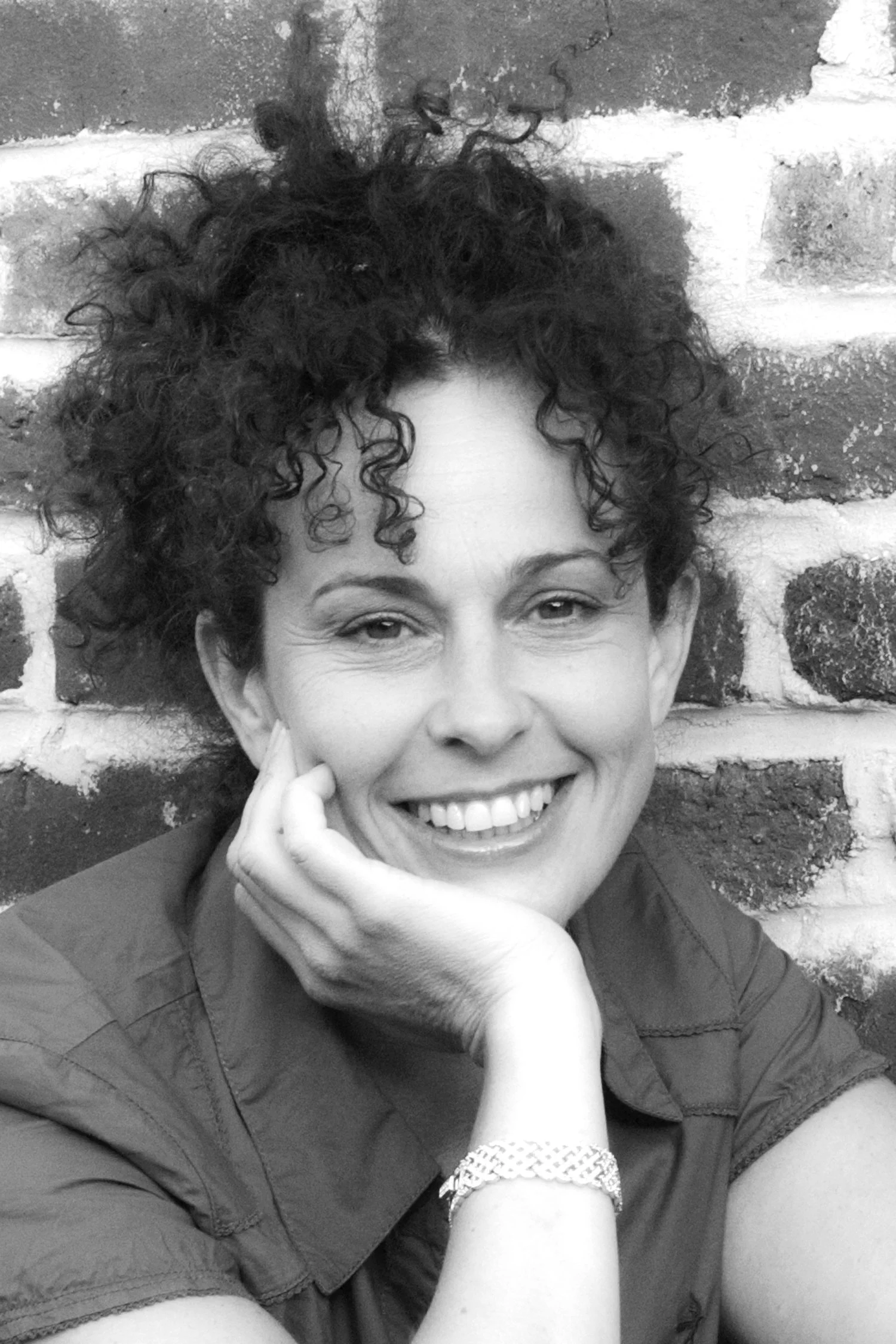 A woman - Justine Claire Photography with curly hair smiling, resting her chin on her hand, in front of a brick wall.