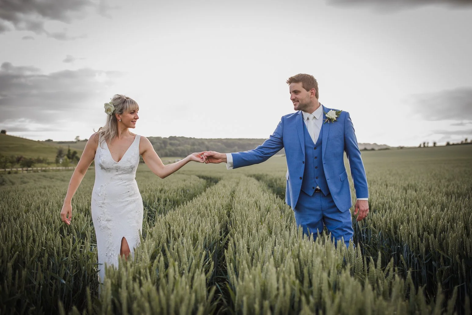 A bride and groom holding hands in a green wheat field with rolling hills in the background, overcast sky, wedding attire.