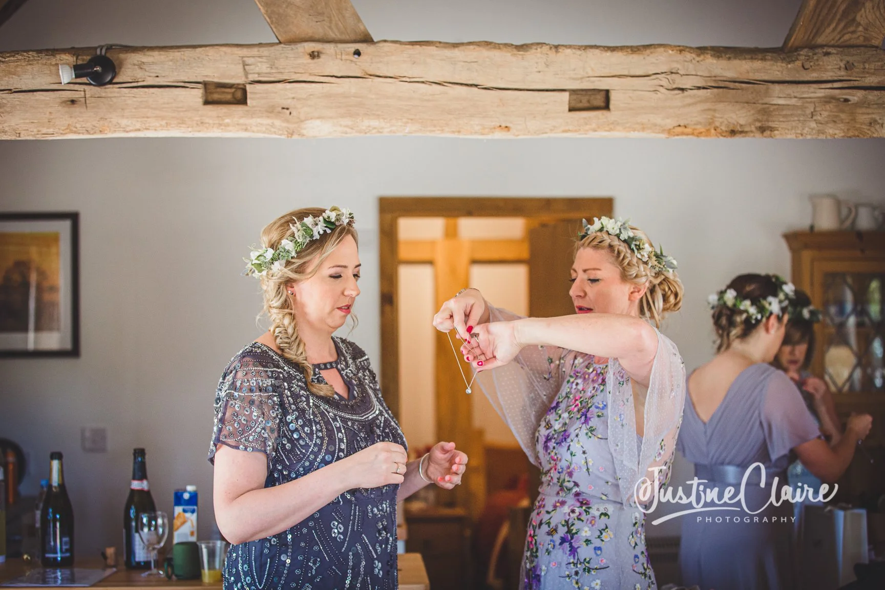 Two women wearing floral crowns preparing for a wedding, one is wearing a dark patterned dress and the other in a pastel floral dress, in a rustic room with wooden beams and furniture.