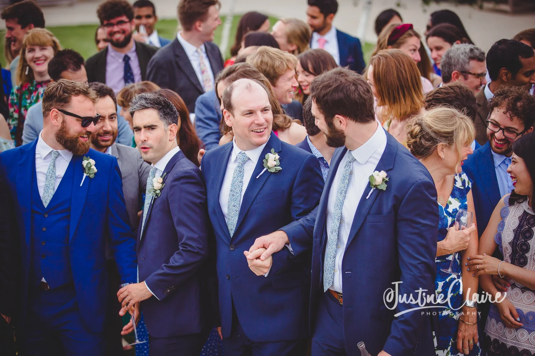 Group of people at a wedding celebration, many dressed in suits with boutonnieres, smiling and engaging in conversation outdoors.