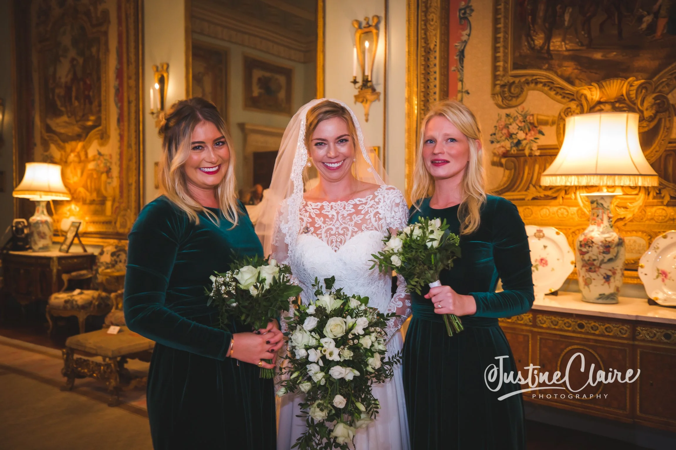 A bride in a white lace wedding gown and veil standing between two bridesmaids in dark green velvet dresses, holding bouquets of white roses, in an ornate room with gold accents and antique lamps.
