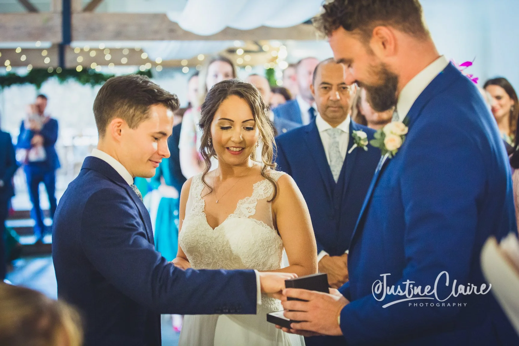 A couple exchange wedding vows while holding a ring box, surrounded by family and friends at a wedding ceremony