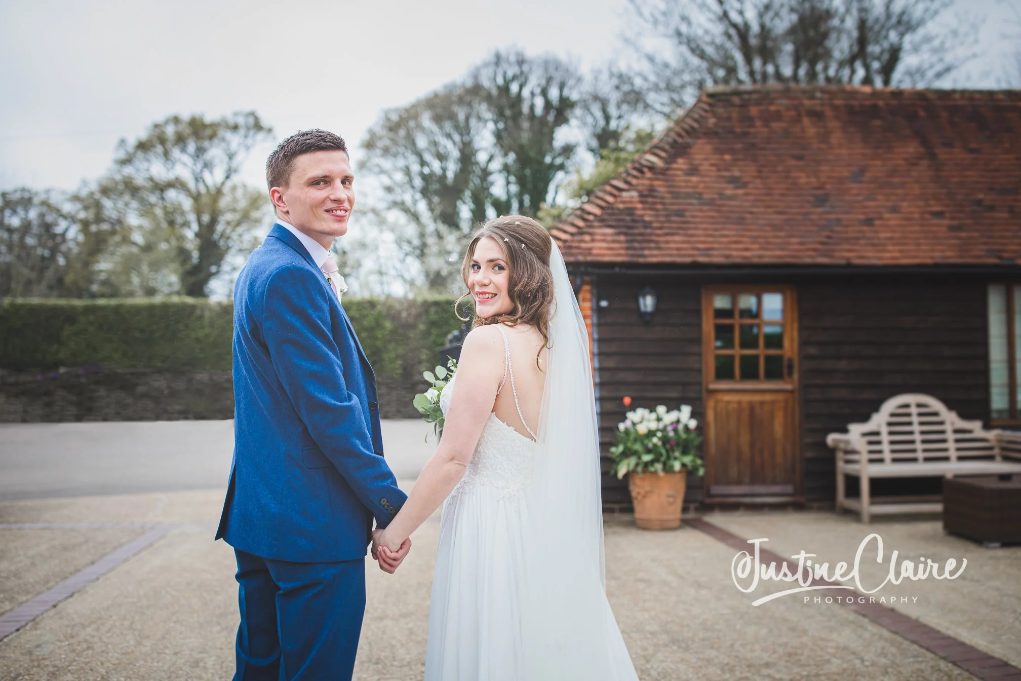 A newlywed couple holding hands outdoors, at Fitzleroi Barn in Fittleworth with the groom in a blue suit and the bride in a white wedding dress, standing in front of a wooden building with a potted flowering plant nearby, during daytime.