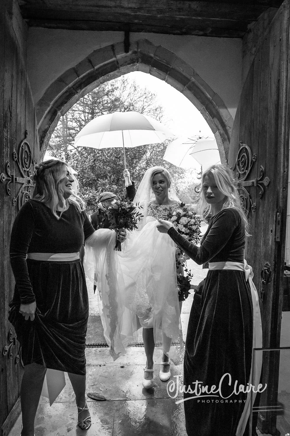 Black and white photo of three women, one in a wedding dress holding a bouquet, standing in a doorway with rain outside, two women in dark dresses with white sashes, one holding a bouquet and the other holding an umbrella.