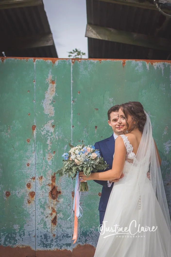 A bride and groom posing for a wedding photo in front of a weathered green wall. The groom is in a navy suit, and the bride is in a white wedding dress with a veil, holding a bouquet of flowers.