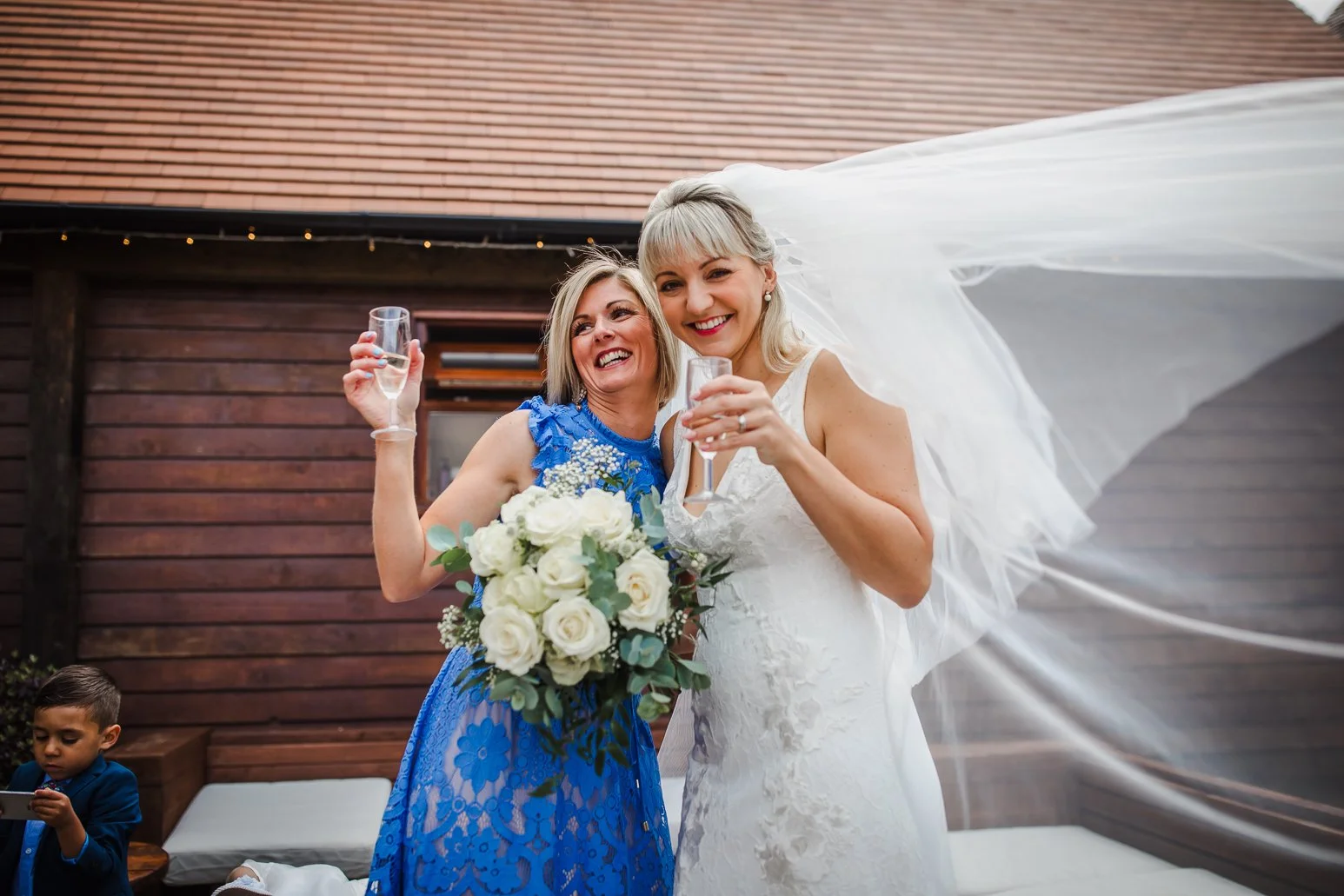 Two women celebrating at a wedding, one in a wedding dress and the other in a blue dress, holding champagne glasses, with a young boy in the background looking at a device.
