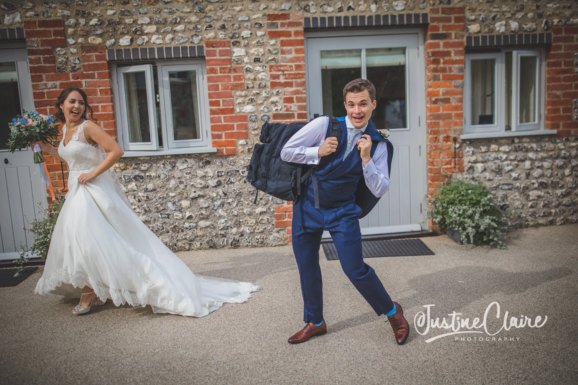 A bride in a white wedding dress and a groom in a blue suit with a backpack outside a brick and stone building, both smiling and appearing excited.