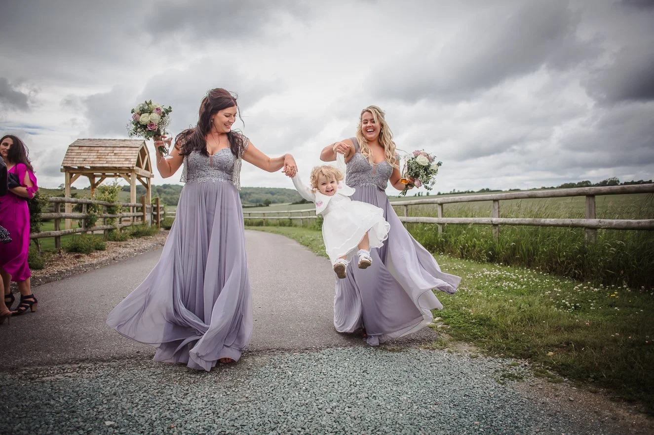 Two women in lavender bridesmaid dresses holding hands with a young girl in a white dress, lifting her while walking on a country road surrounded by grass and a wooden fence, with cloudy skies overhead.