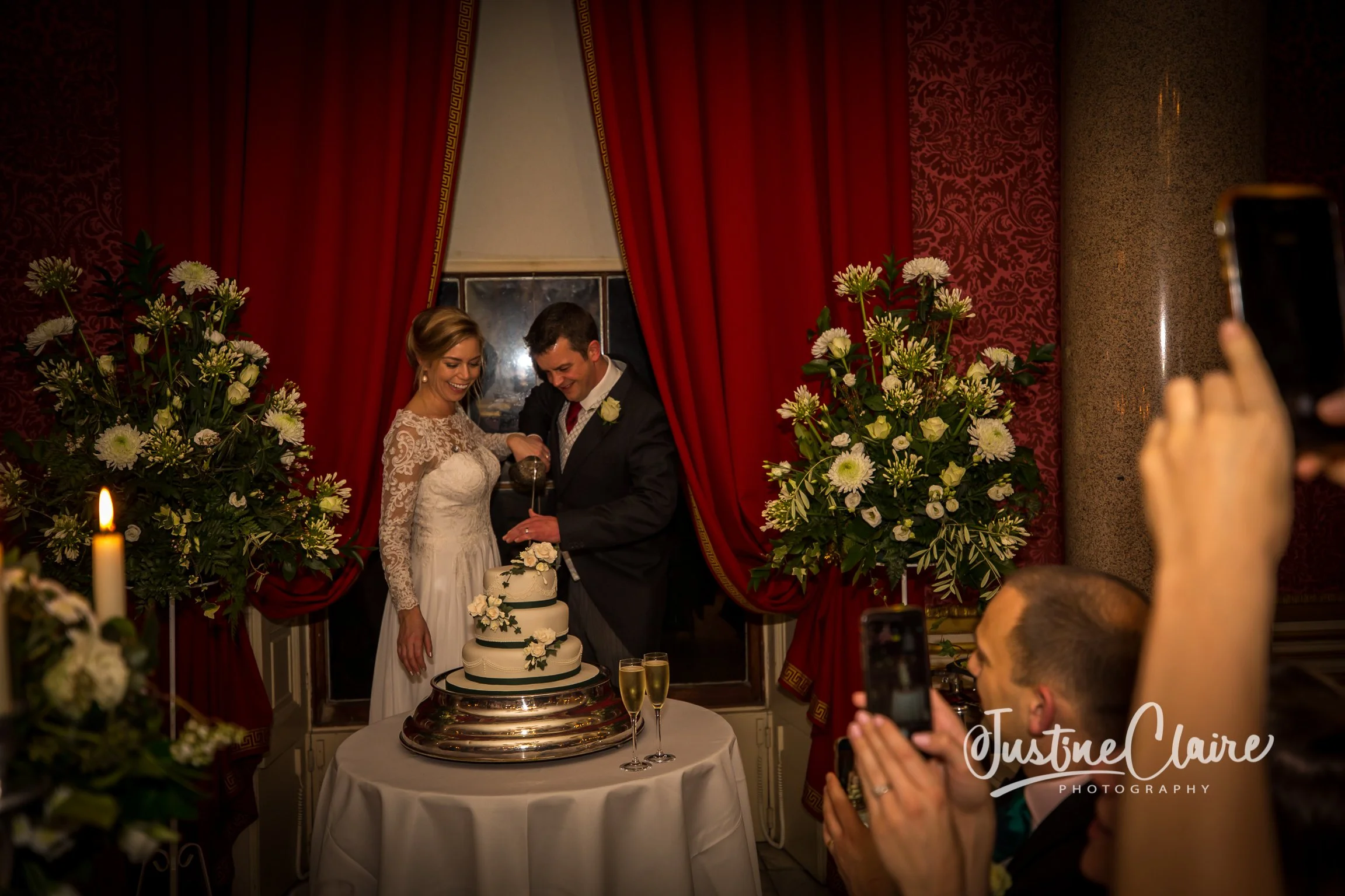 Couple in wedding attire cutting a wedding cake surrounded by large floral arrangements, with guests taking photos, in an elegant decorated room with red curtains.