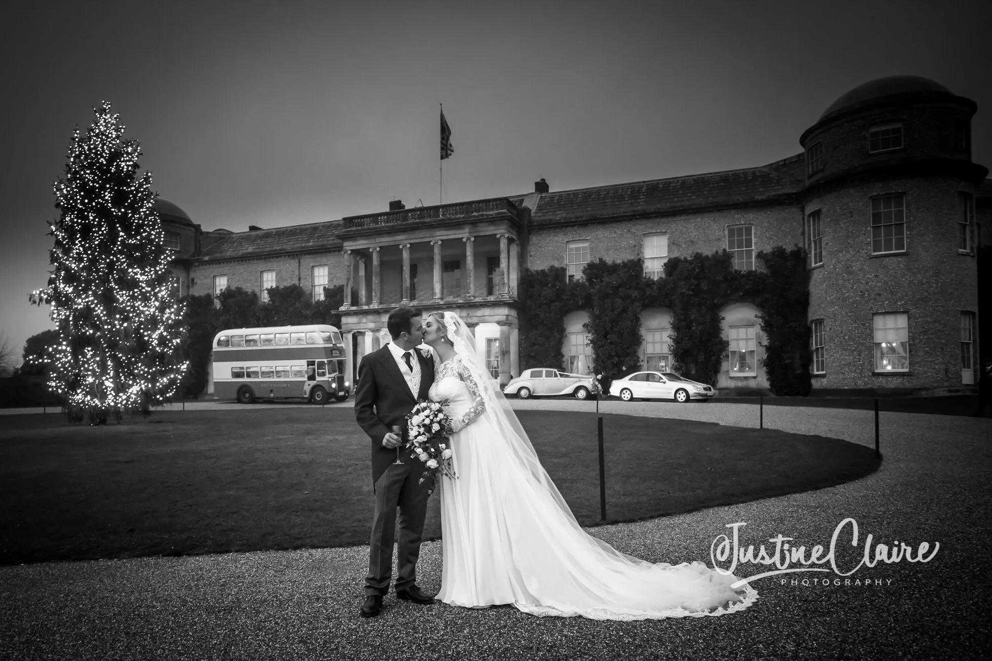 A black and white photo of a bride and groom kissing in front of a large, historic building. The bride wears a long white gown with lace sleeves and a veil, holding a bouquet, while the groom wears a suit and tie. A decorated Christmas tree is on the