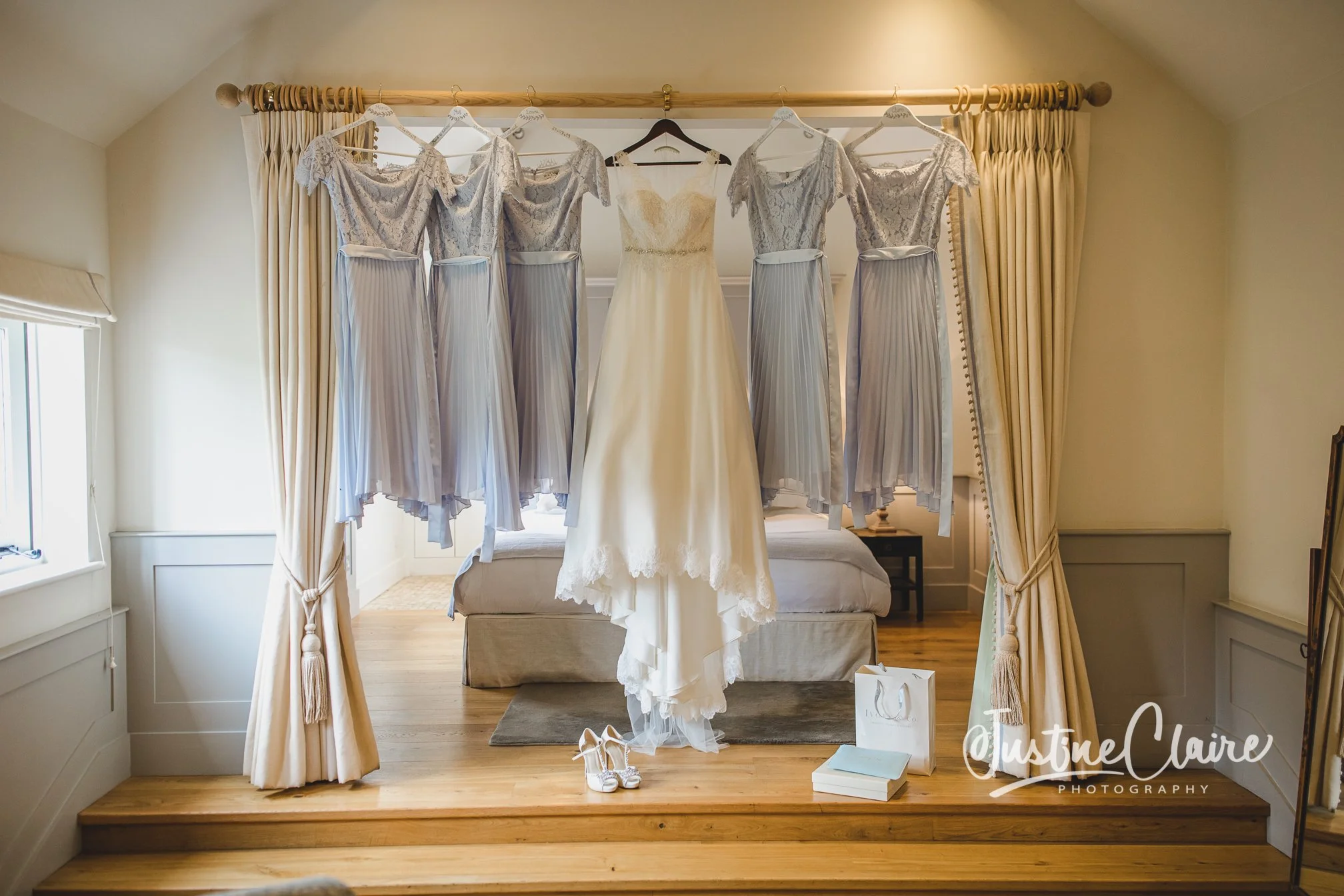 A wedding dress hanging on a rack with four bridesmaid dresses, a pair of white high heels, a gift bag, and a book on a wooden platform inside a room with beige walls and cream curtains.