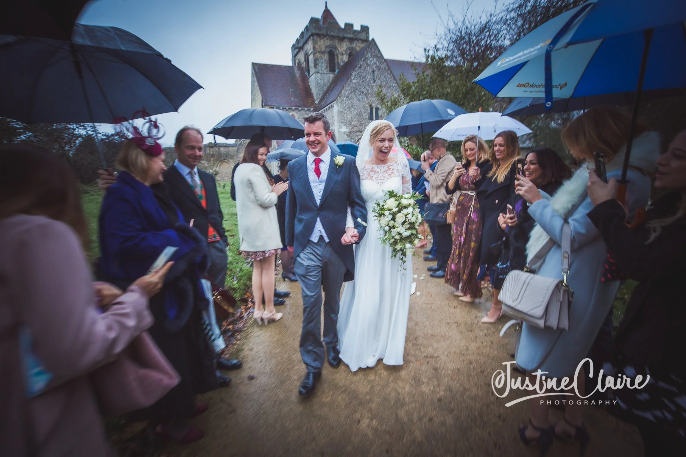 Bride and groom walking arm-in-arm on a rainy day, surrounded by guests with umbrellas, near a church, celebrating their wedding.