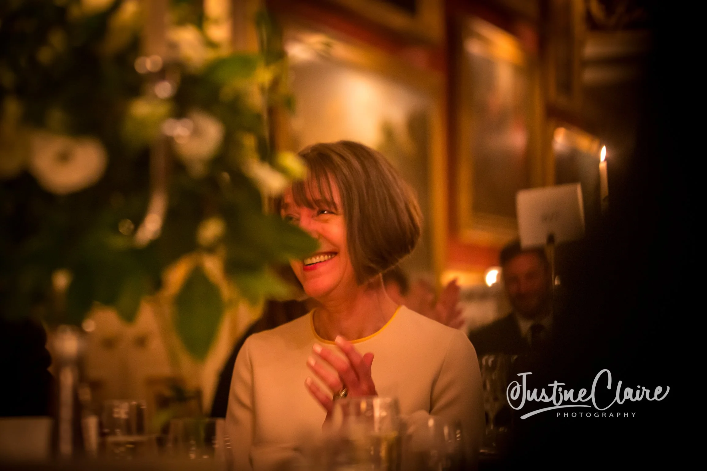 A woman with short brown hair smiling and clapping her hands at a dinner event. She is wearing a cream-colored top with yellow trim. The background includes warm lighting, framed pictures, and other people dining.