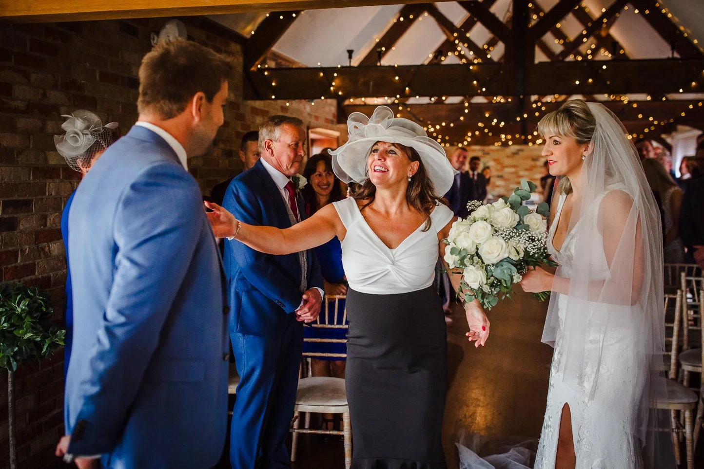A wedding reception scene with a woman in a large white hat smiling and reaching out to a man in a light blue suit. A bride holding a bouquet of white roses looks on. Guests are in the background under string lights in a rustic venue.