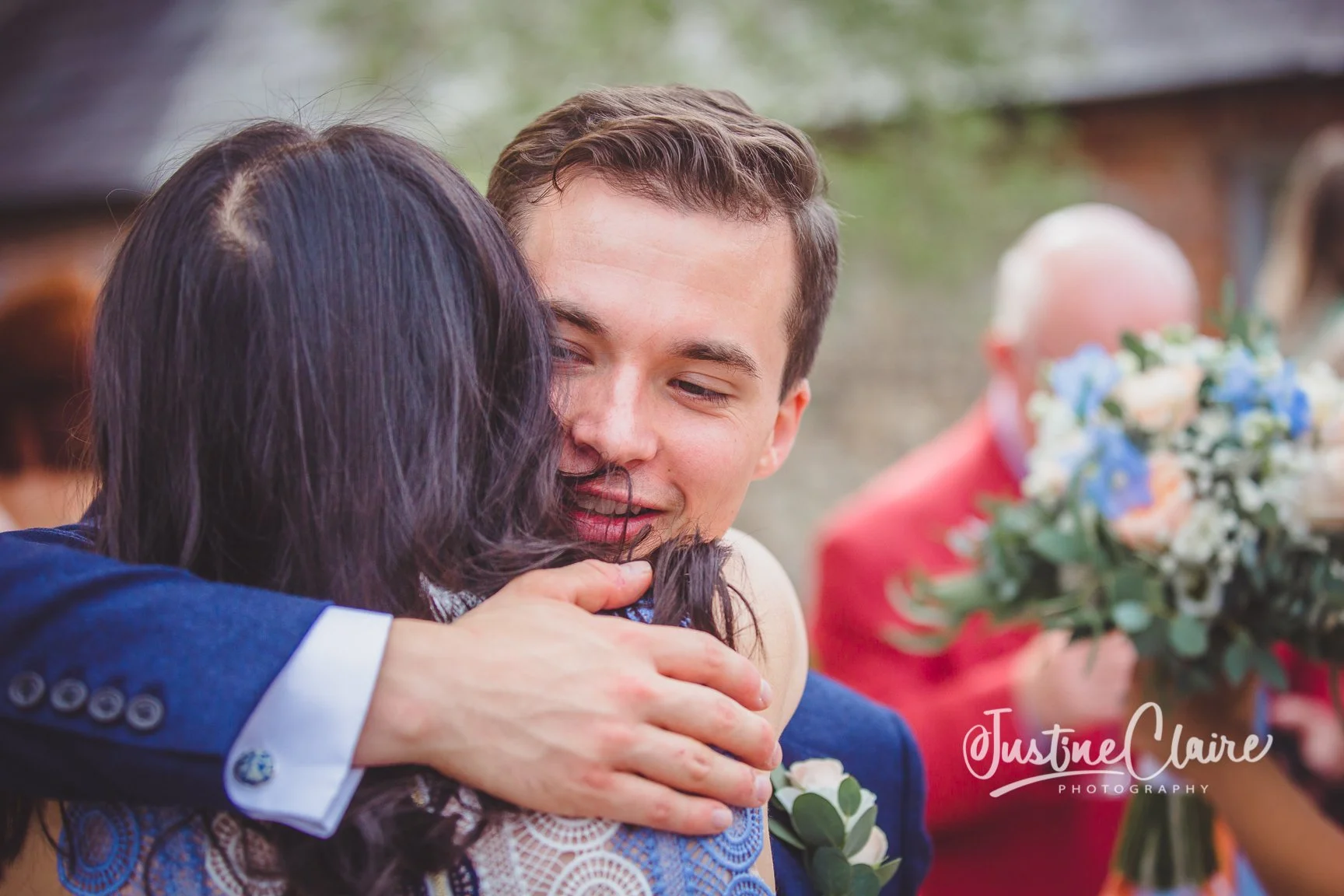 A young man and a woman are hugging at a wedding, with the woman’s back to the camera and the man smiling partially visible. In the background, an older man is holding a bouquet of flowers. candid photography
