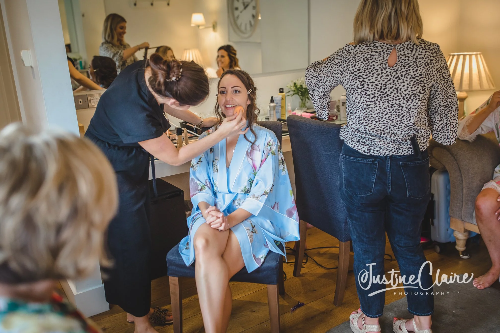 A woman in a floral silk robe is getting her makeup done by a makeup artist in a room with mirrors and a clock on the wall. Other women are getting ready, and there are beauty products on the counter.