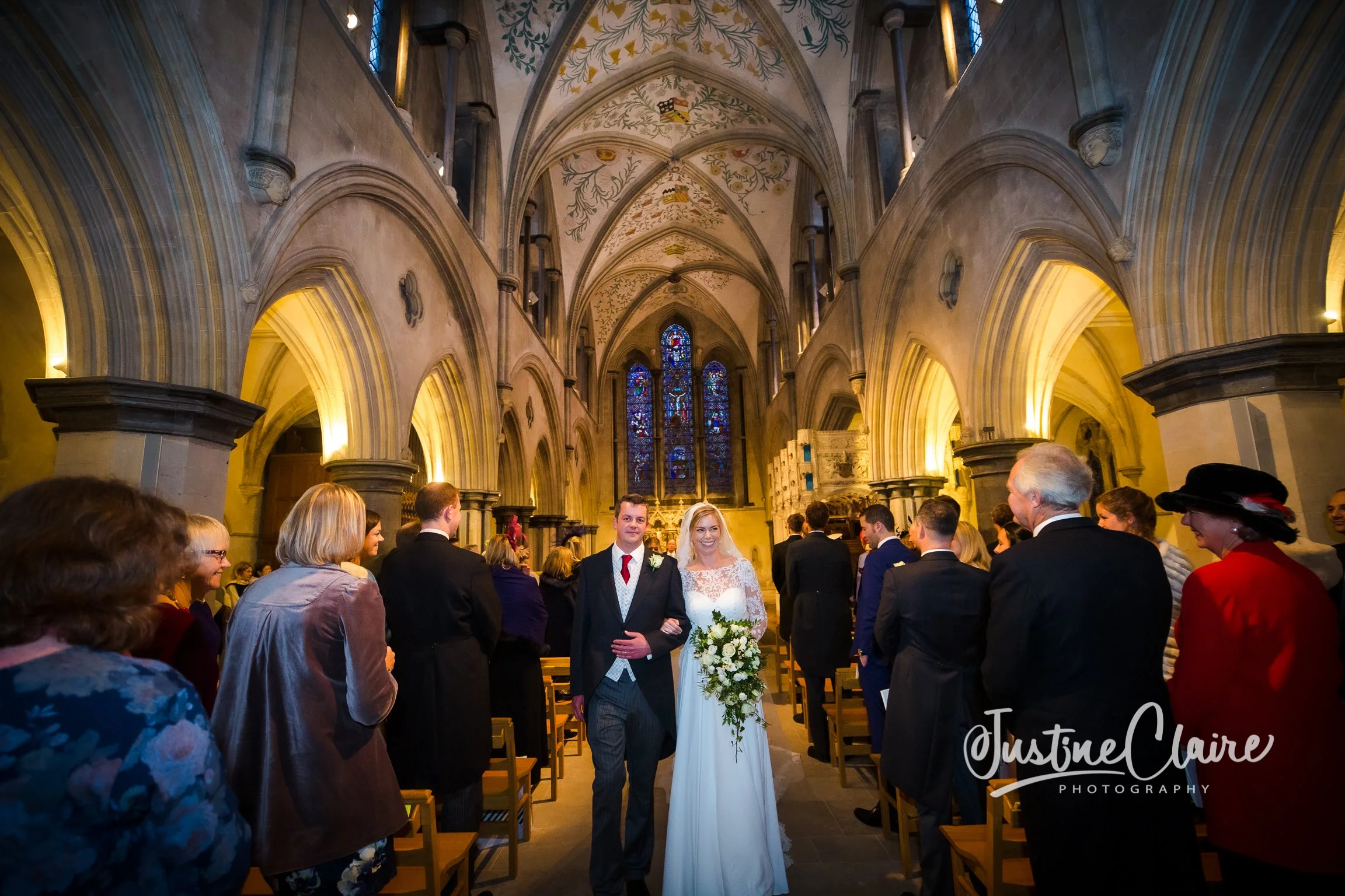 A bride and groom walking down the aisle in a Gothic-style church with guests on either side, stained glass windows at the altar, and ornate stone arches overhead.