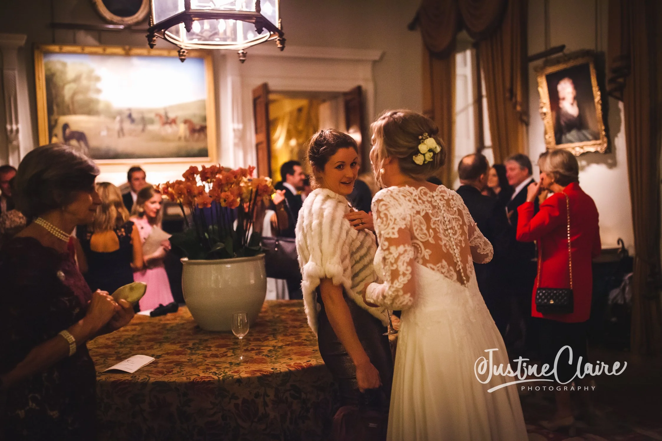 Wedding reception with guests socializing indoors, a floral arrangement on a table, and a bride in a lace dress with flowers in her hair.