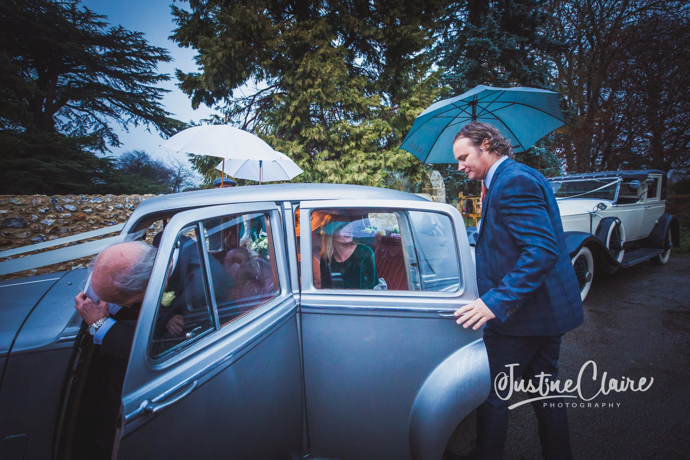 A man in a blue suit opening the door of a vintage silver car with a woman inside, and another man adjusting his glasses outside the car during a rainy day, with trees and an old stone wall in the background.