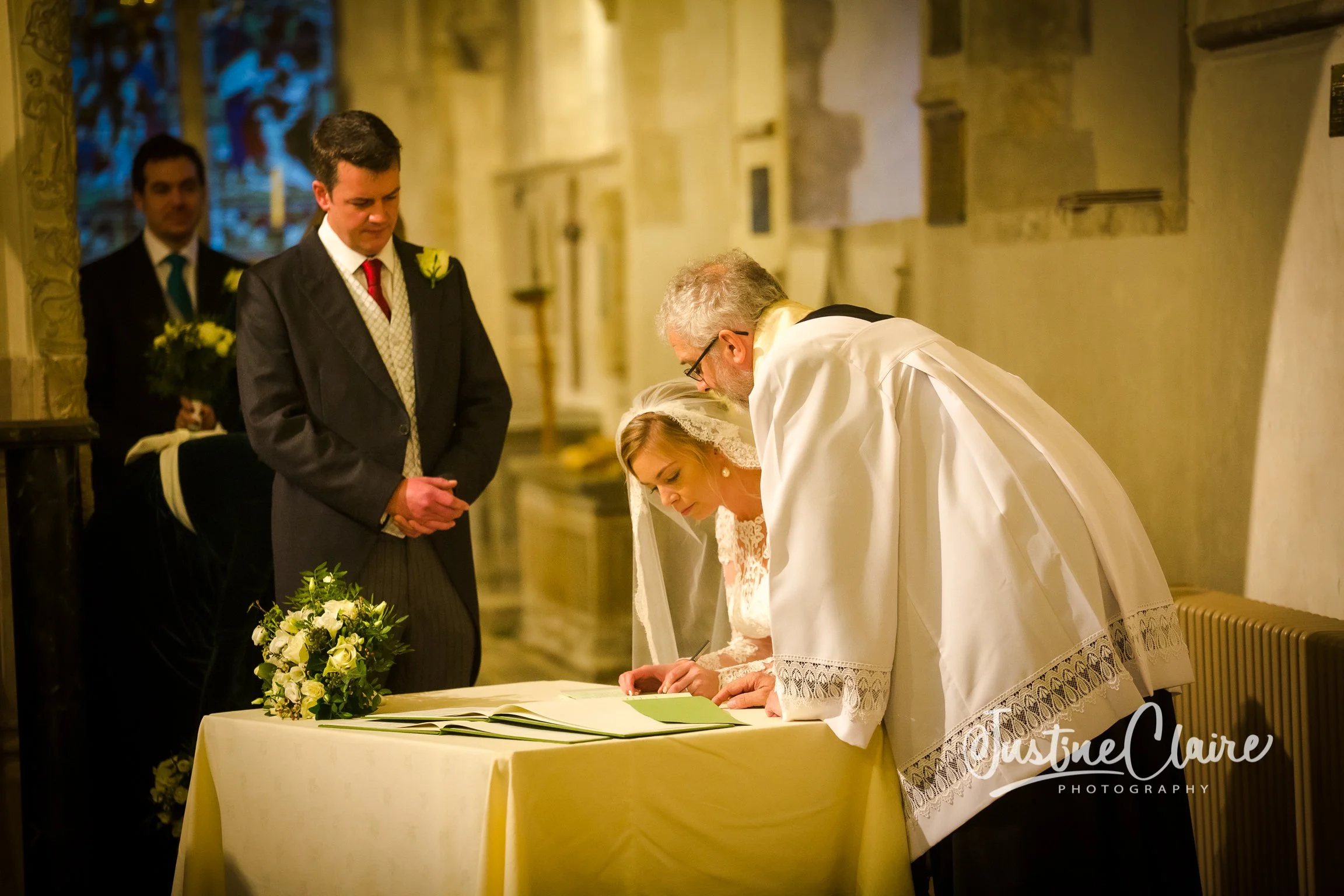 A wedding ceremony in a church with a bride signing a document, accompanied by a priest and a groom, with two groomsmen standing nearby.