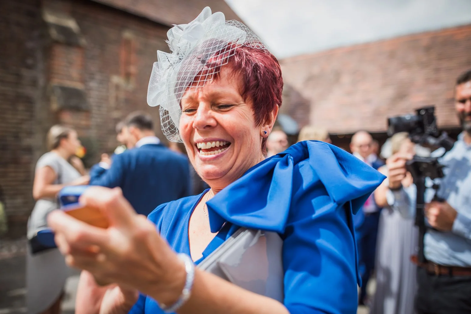 A smiling woman with red hair, wearing a blue dress and a white fascinator, looks at her phone during an outdoor celebration with people and cameras in the background.