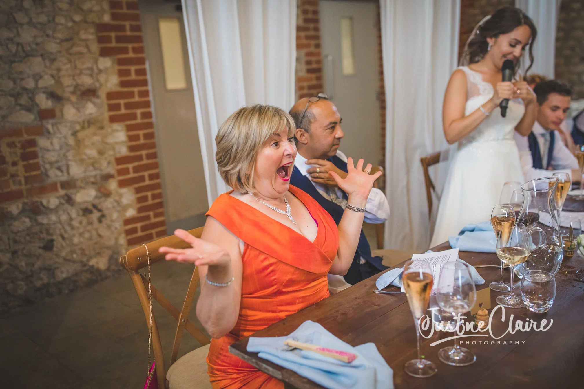 Woman wearing an orange dress with short blonde hair, smiling and gesturing with her hands during a reception, with a woman in a wedding dress speaking into a microphone in the background.